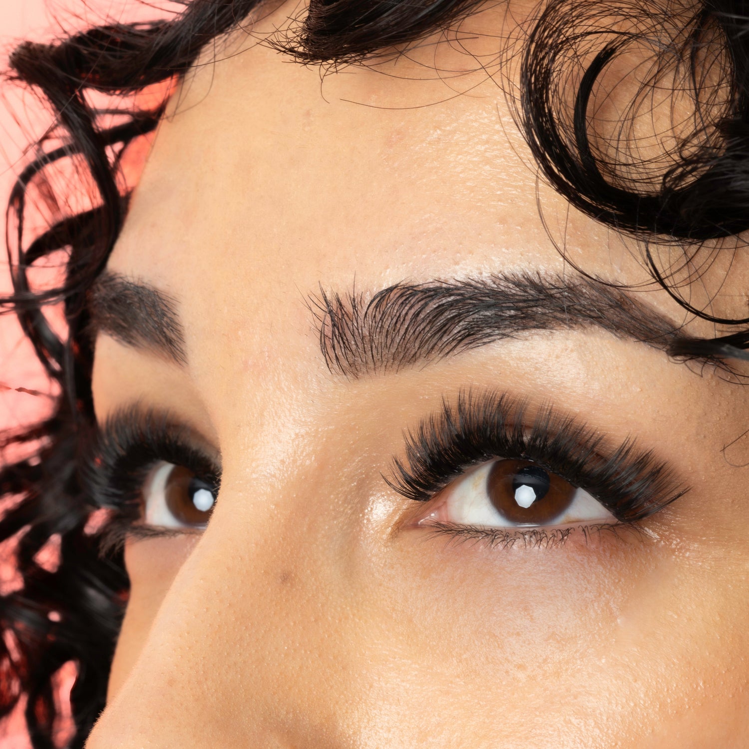 Close-up of a person's eye area showcasing long, dark, curled eyelashes against a solid peach-pink background.