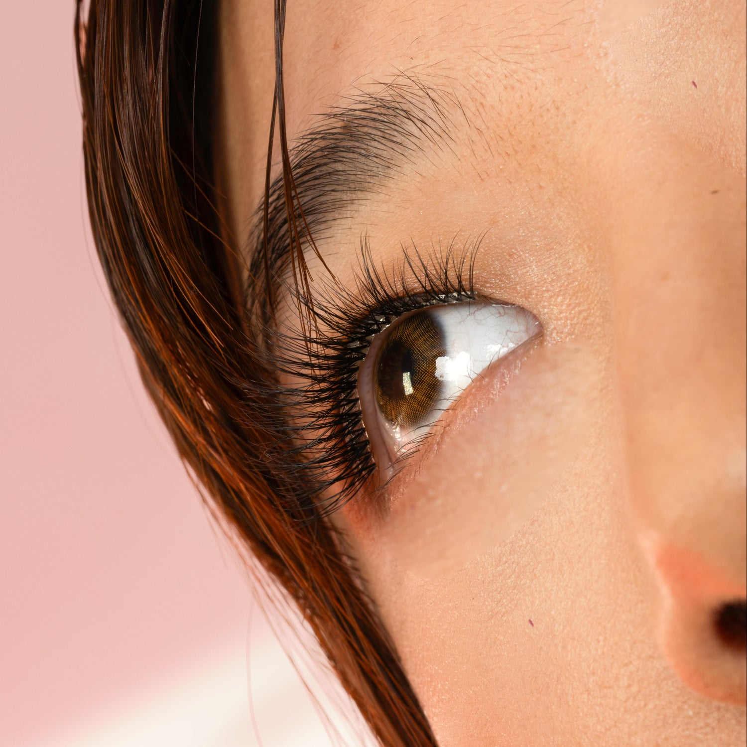 Close-up of a person's eye with long dark eyelashes, brown hair, and a soft pink background.