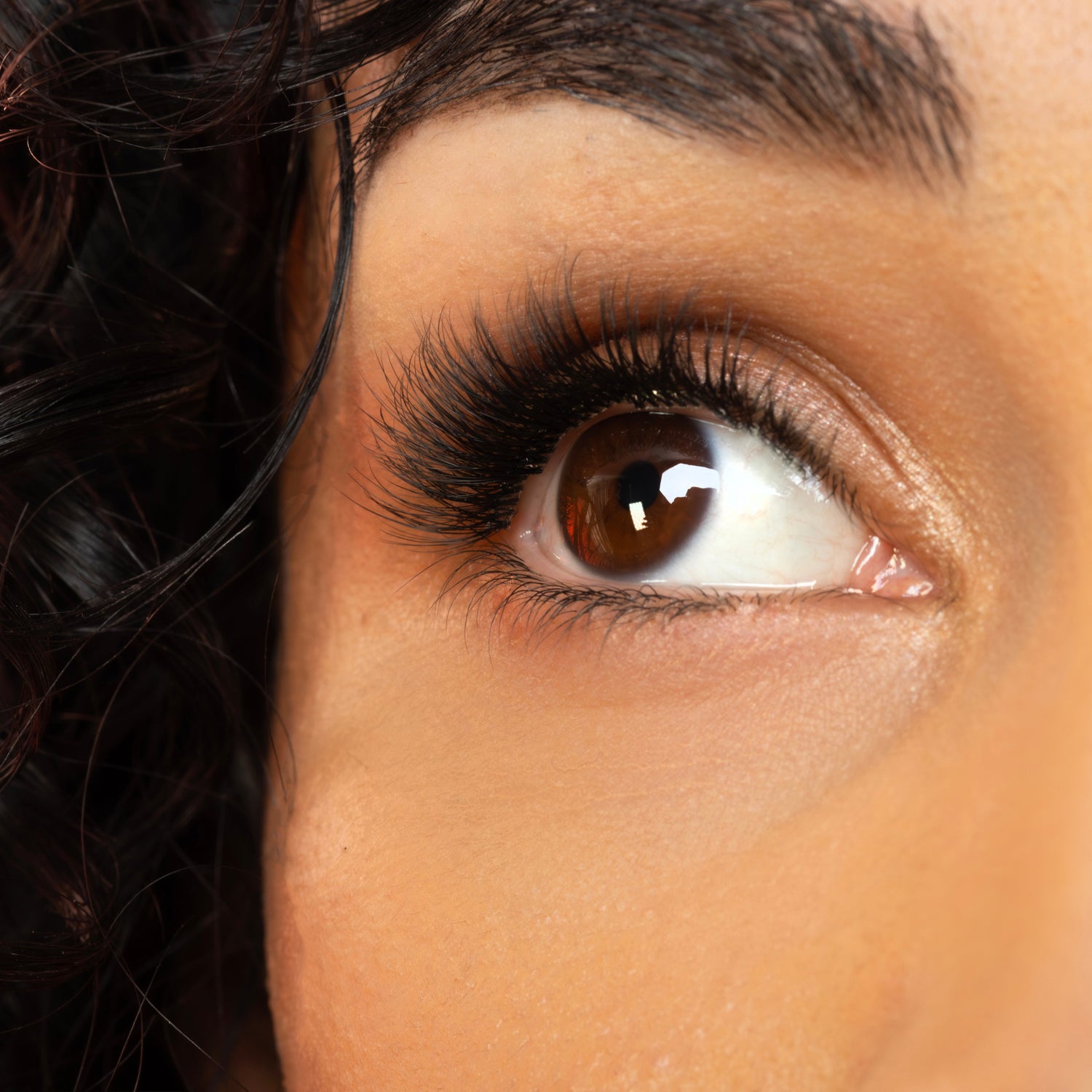Extreme close-up of a person's eye with long black eyelashes, brown iris, dark curly hair, and warm skin tones against a soft pink background.