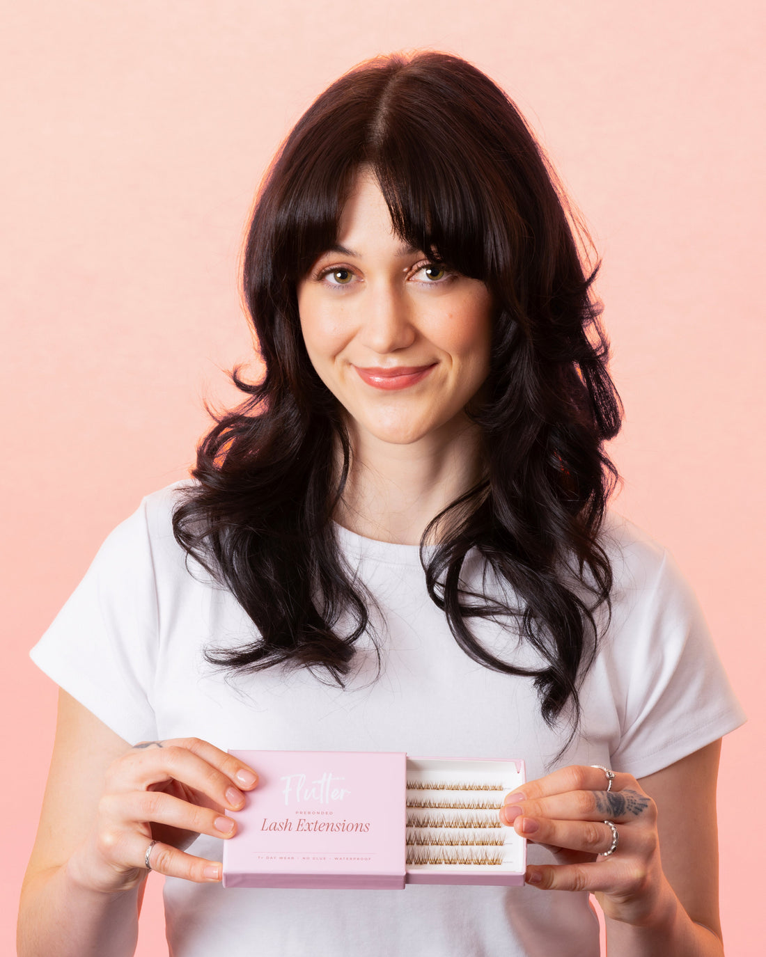 A woman with dark hair and bangs, wearing a white t-shirt, holds a pink box of eyelash extensions against a solid pink background.