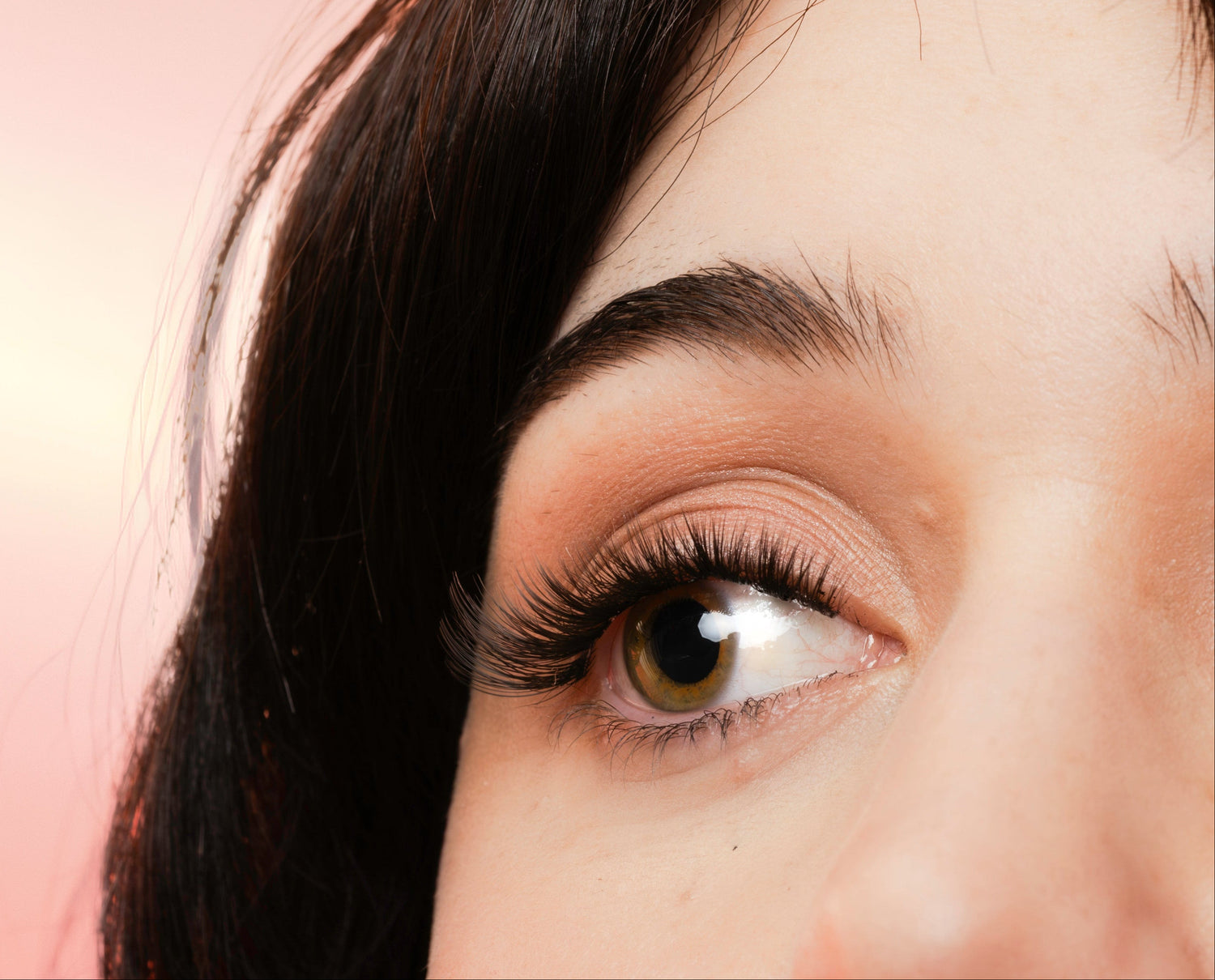 Close-up of a woman's face with dark hair, brown eyes, false lashes and subtle eyeshadow, set against a soft pink background.