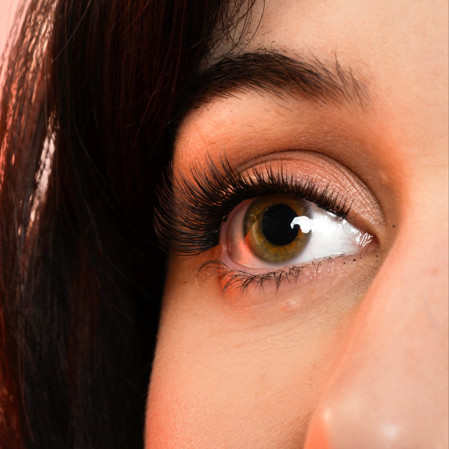 Close-up of a woman's eye wearing long, dark false eyelashes against a soft peach-pink background.